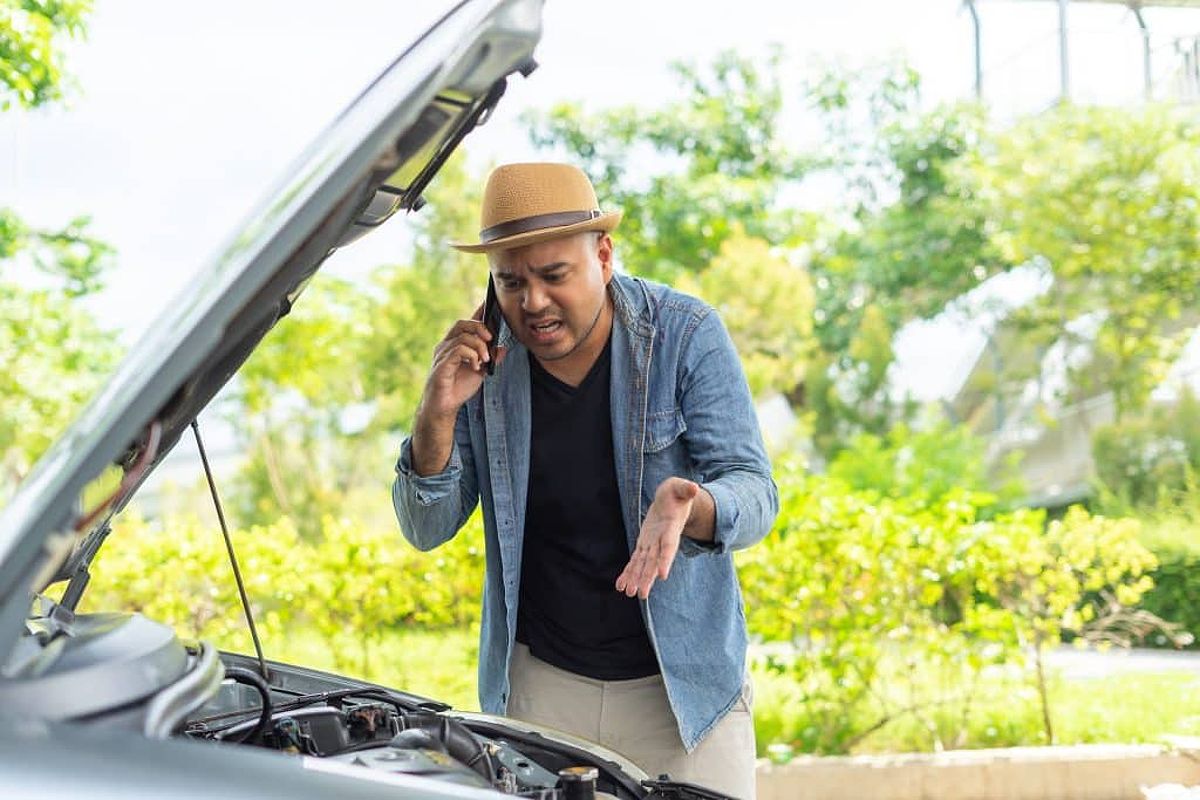 Frustrated young man looking down at car engine while on the phone.